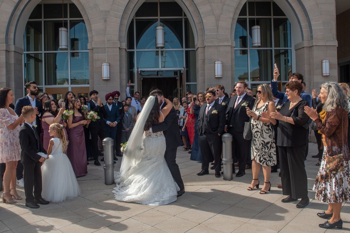 A bride and groom kiss outside a grand stone building as cheering guests with phones capture the moment in warm sunlight.