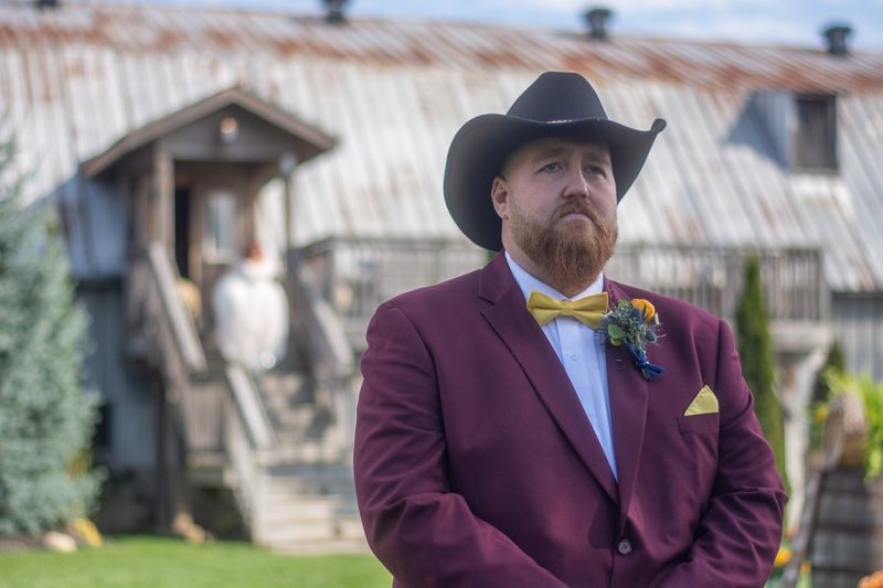 A groom in a maroon suit and yellow bow tie stands by a blurred rustic building as his bride ascends steps.