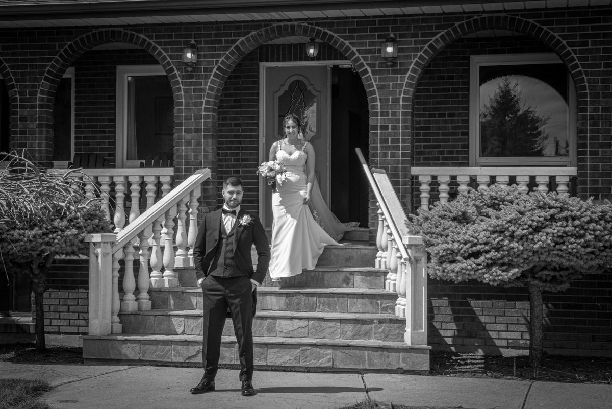A bride and groom stand at the entrance of a brick home, the bride on the porch steps with a bouquet.