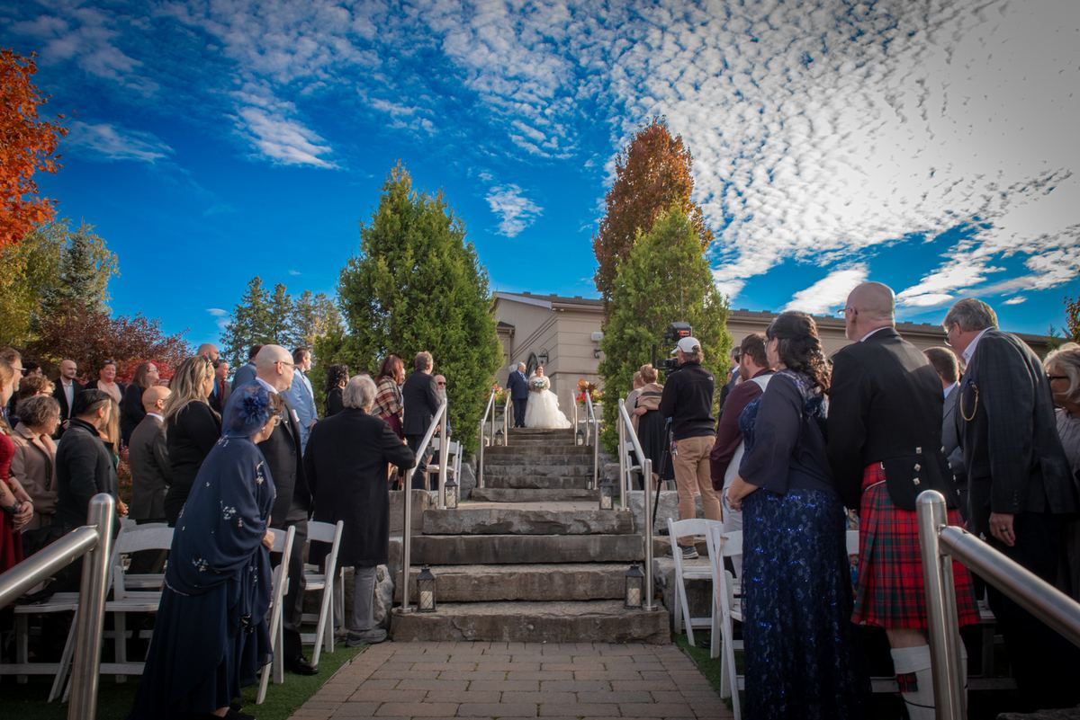 Bride and groom ascend stone steps toward altar under blue sky, surrounded by guests in formal attire.