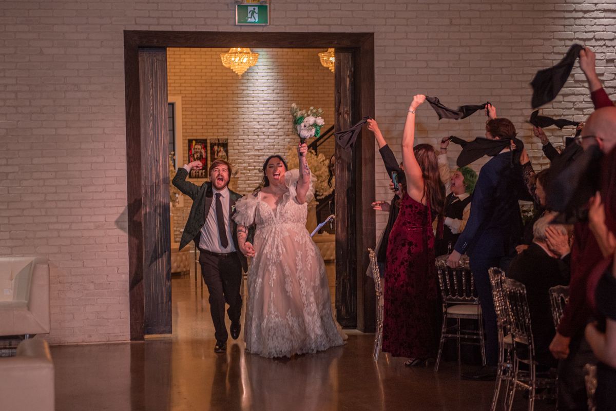 A bride and groom joyfully enter a reception as guests toss jackets in the air, celebrating with warmth and enthusiasm.