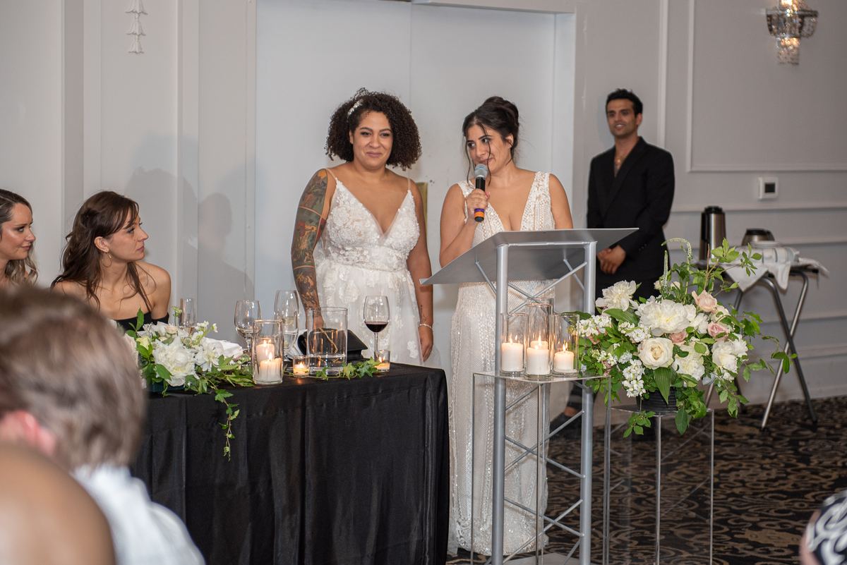 Two brides smiling at a microphone and guests during their wedding reception, surrounded by candles and flowers.