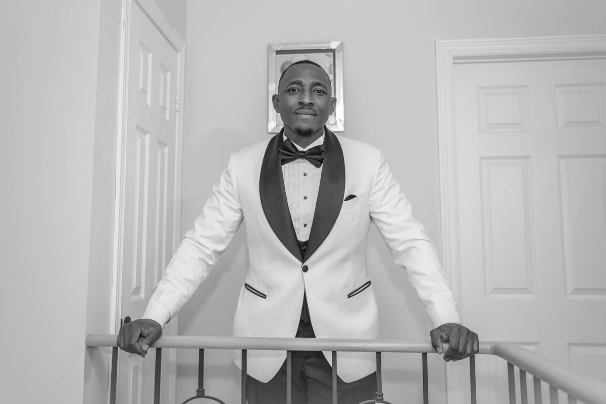 A man in a white tuxedo stands confidently on a staircase railing, looking at the camera during a formal event.
