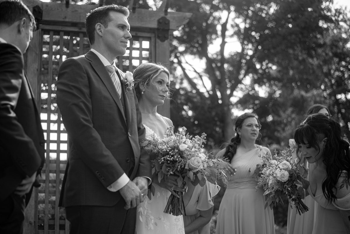 Bride and groom stand beside each other outdoors during their wedding ceremony under dappled sunlight.