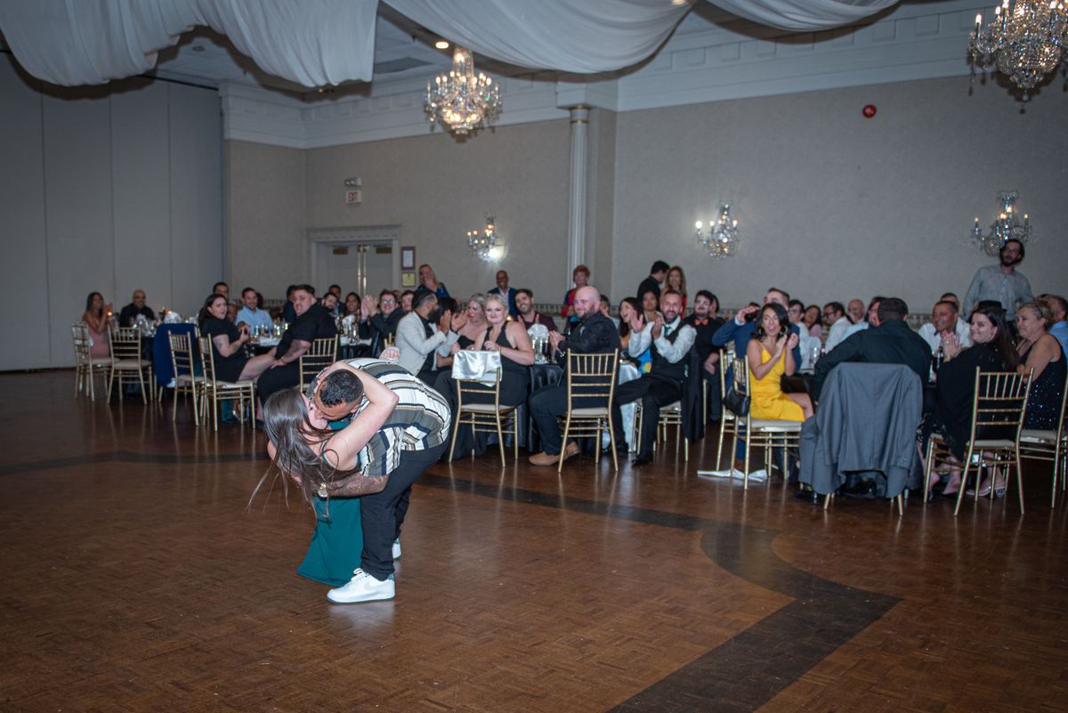 A couple dances intimately in a grand ballroom, performing a dramatic dip as guests applaud and chandeliers cast warm light.