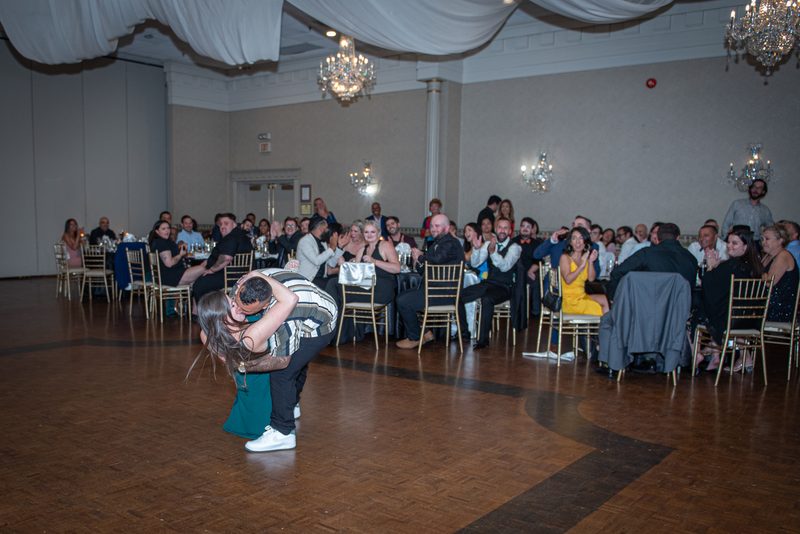 A couple dances intimately in a grand ballroom, performing a dramatic dip as guests applaud and chandeliers cast warm light.