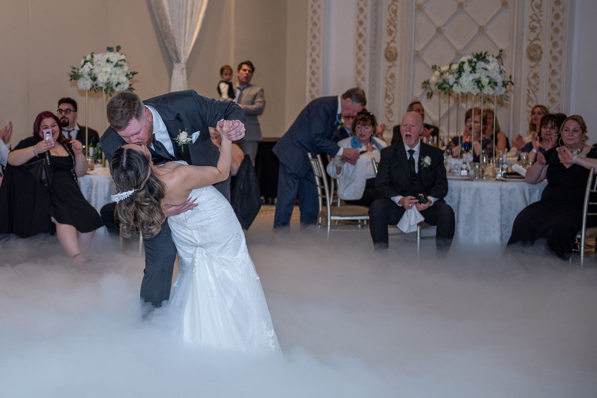A bride and groom dance intimately in foggy light as guests watch from shadowed tables, celebrating their first dance.
