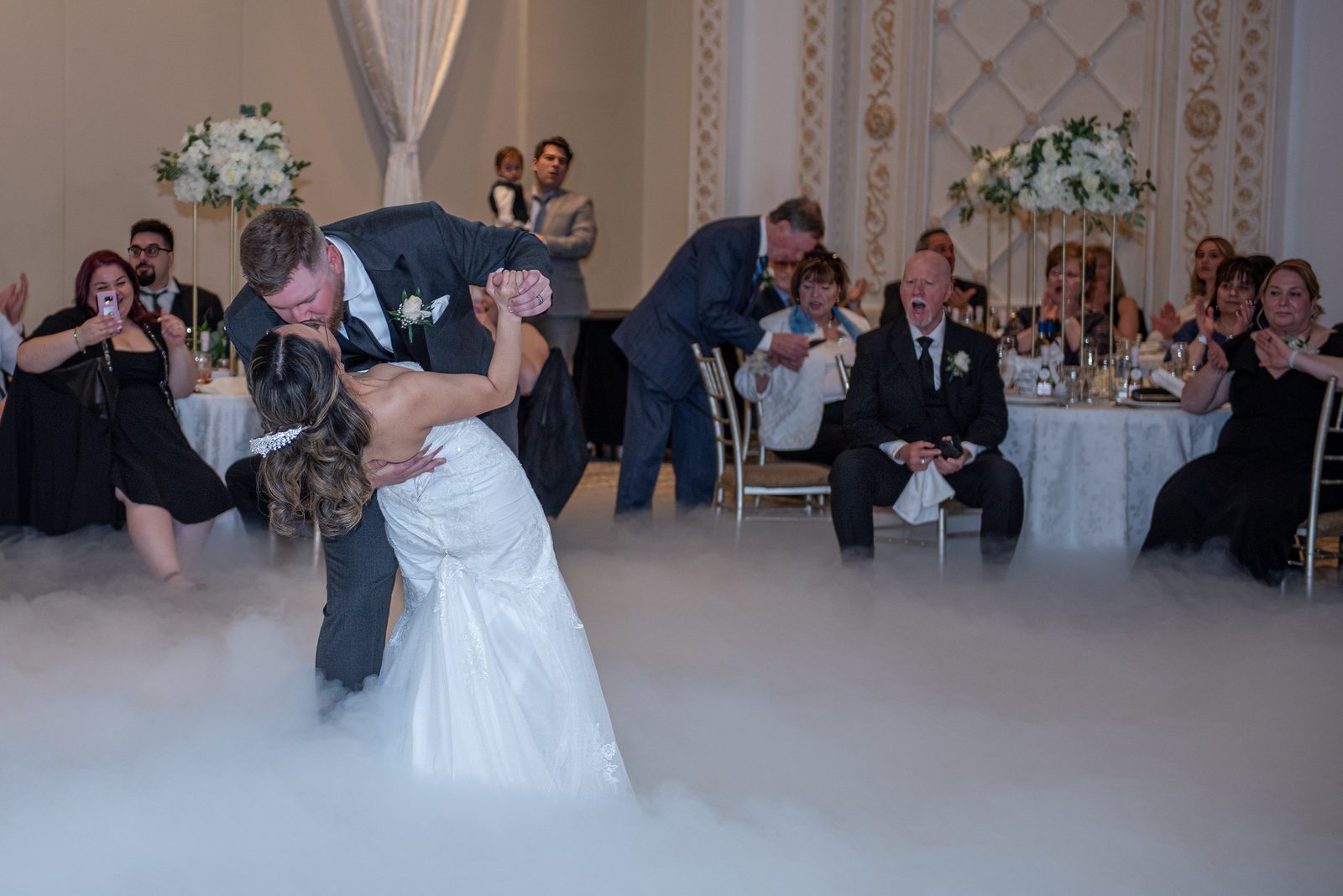 A bride and groom dance intimately in foggy light as guests watch from shadowed tables, celebrating their first dance.