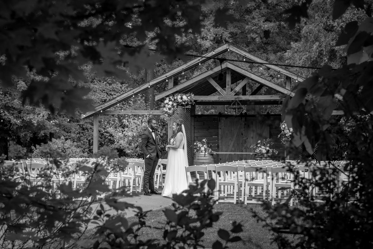 A bride and groom stand at an altar under a wooden structure, surrounded by foliage and empty chairs, in soft light.