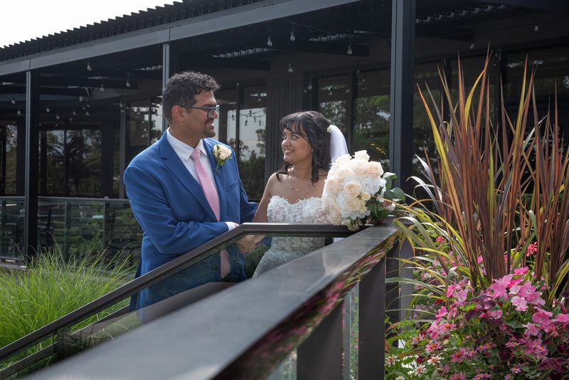 A smiling bride in a white gown and a groom in a blue suit stand on a balcony, gazing at each other amidst greenery.