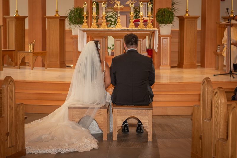 A bride and groom sit facing the altar in a warmly lit church, their formal attire standing out against the sacred setting.