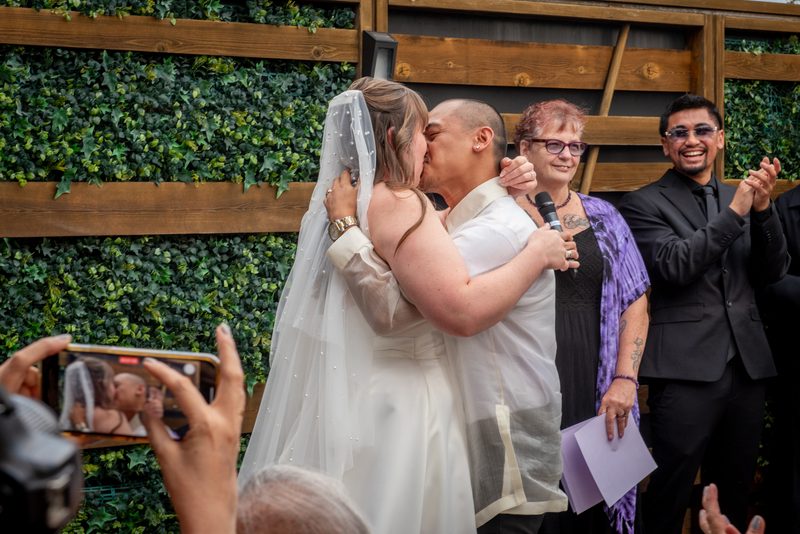 A bride and groom kiss joyfully outdoors, surrounded by clapping guests and a green ivy-covered backdrop.