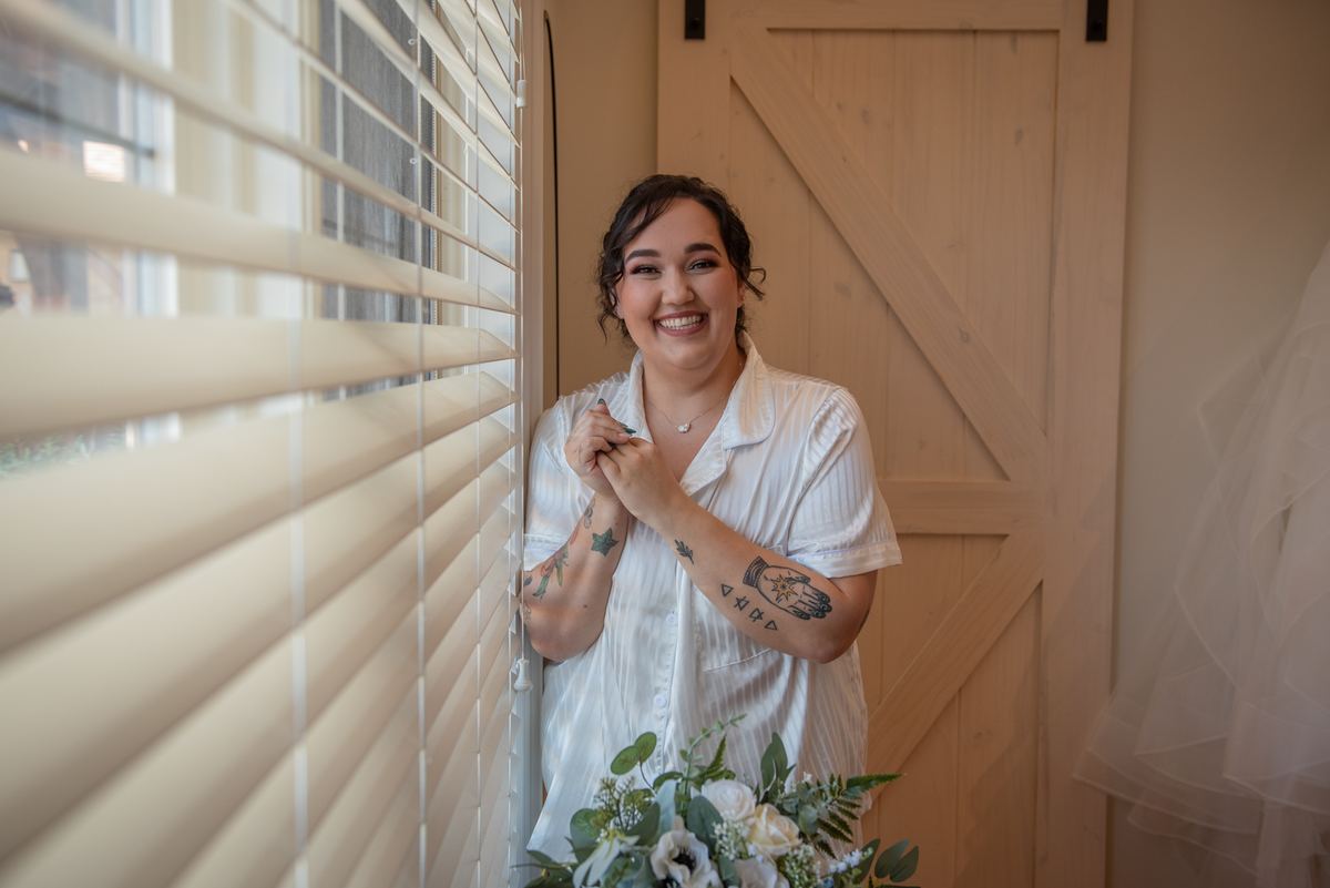 A smiling bride in a white shirt with tattoos peeking through blinds, holding flowers and a veil near a wooden door.