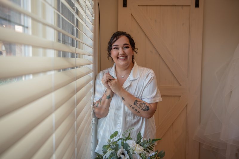 A smiling bride in a white shirt with tattoos peeking through blinds, holding flowers and a veil near a wooden door.