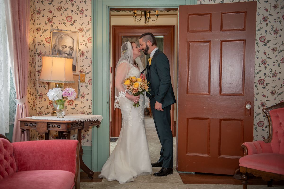 A bride and groom kiss in a vintage room with floral wallpaper and pink velvet chairs, framed by an open wooden door.