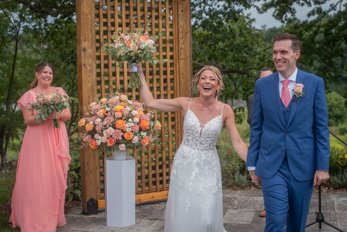 Bride in white lace laughs with bouquet aloft as groom and bridesmaid walk beside her down stone aisle.