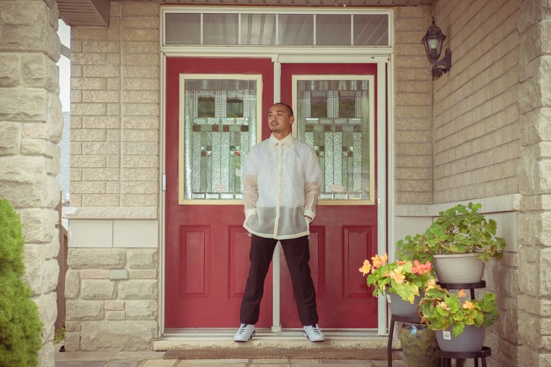 A man in a white blouse stands calmly on porch steps before red double doors, gazing thoughtfully at the sky.