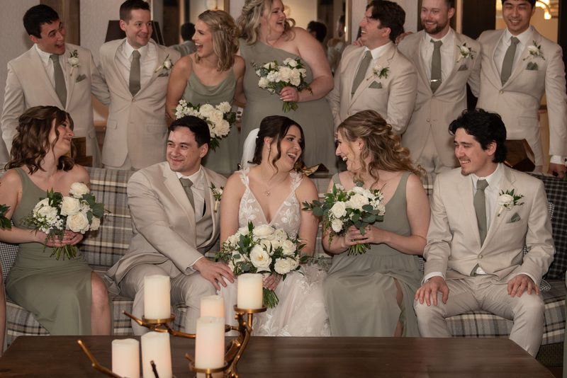 A joyful wedding party, including the bride and groom, gathers around a candlelit table, exuding warmth and camaraderie.