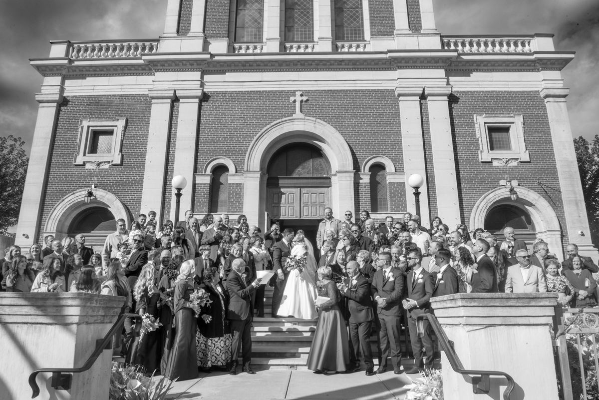 A bride and groom stand with guests on the steps of a grand brick church, bathed in sunlight with sharp shadows.