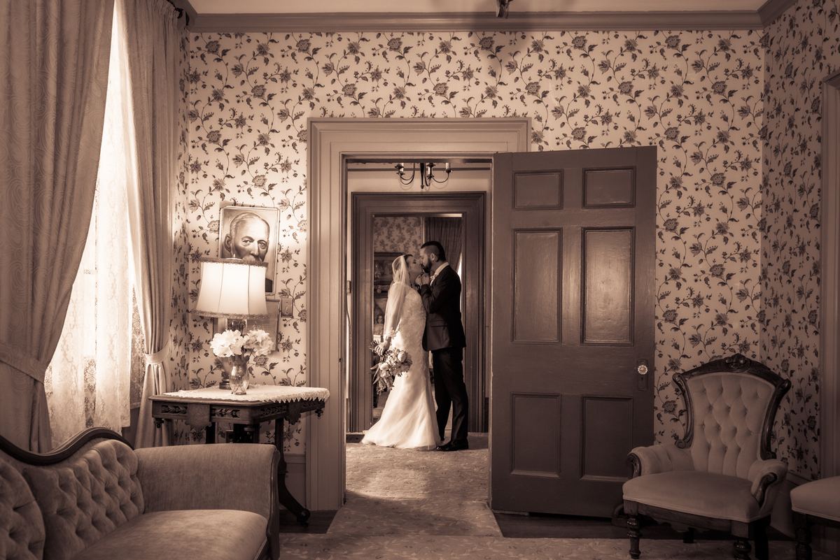 A newlywed couple kisses in a vintage doorway with floral wallpaper and antique furnishings, illuminated by soft light.