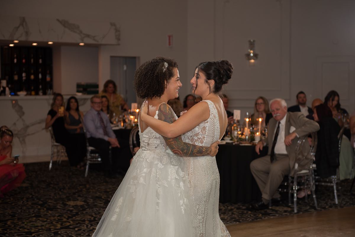 Two brides dance joyfully together at their wedding, surrounded by smiling guests and candlelit tables.