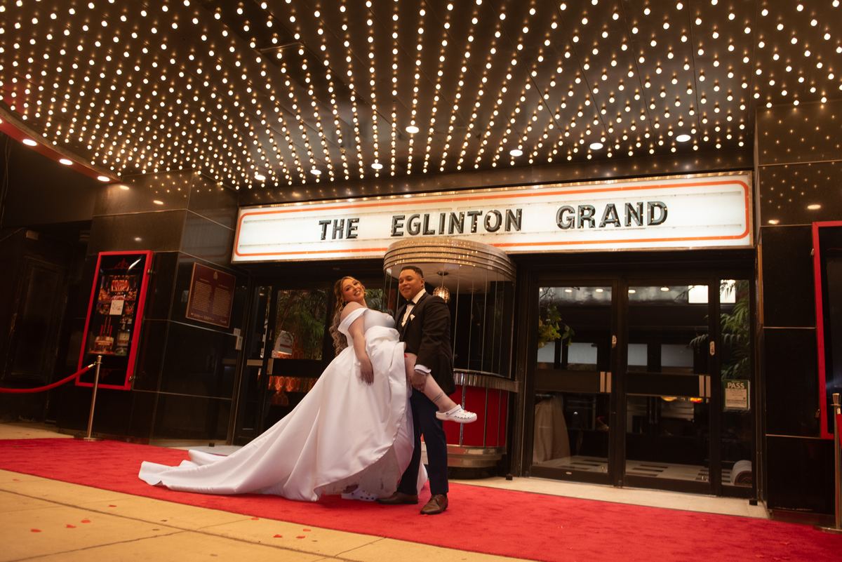 A bride and groom smile joyfully, lifting her trailing gown on a red carpet adorned with twinkling lights.