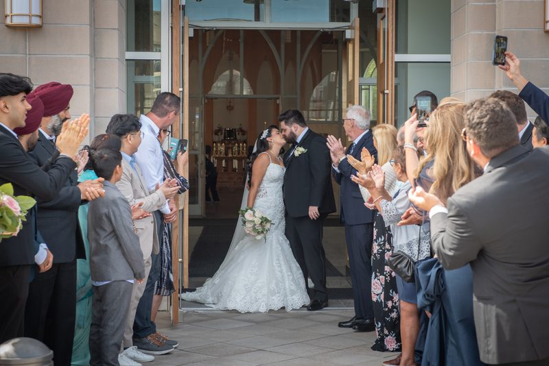 A newlywed couple exits a church, sharing a joyful kiss as guests applaud and capture the moment on their phones.