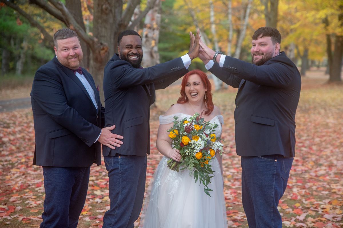 A laughing bride in a white gown surrounded by playful groomsmen high-fiving, set against autumn foliage.
