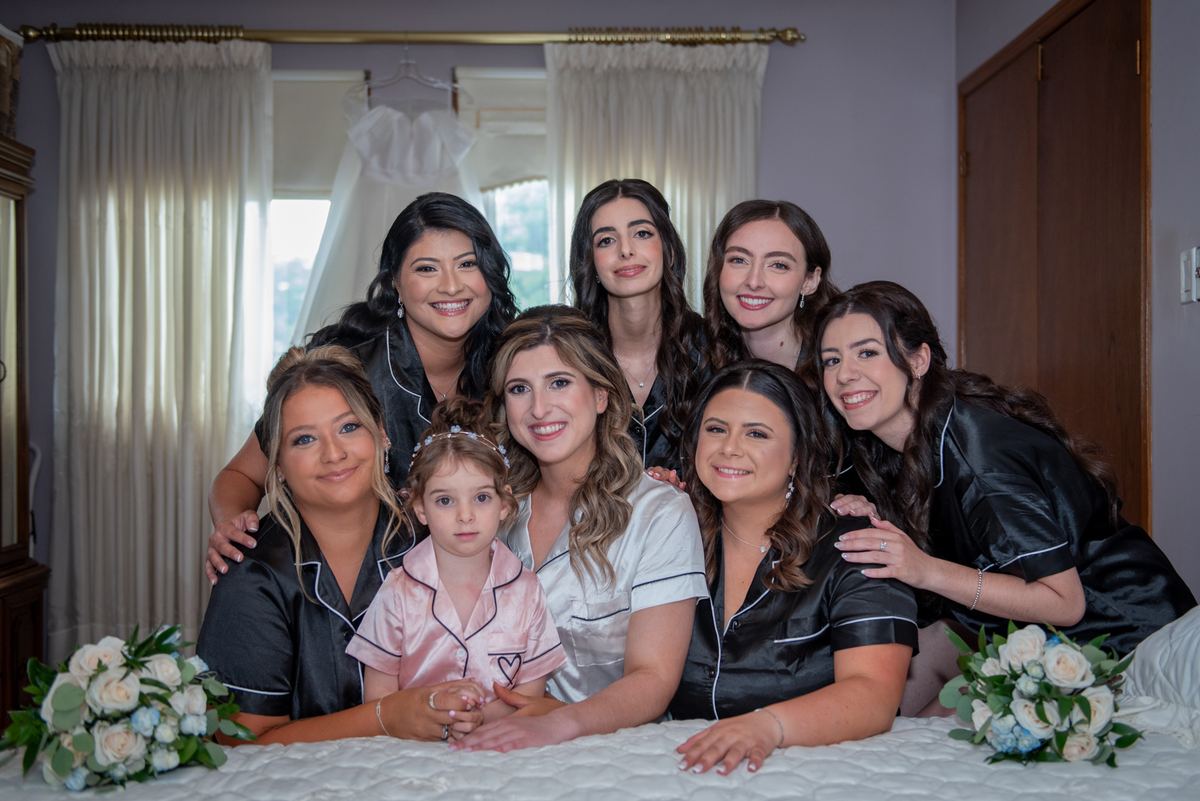 A bride, bridesmaids, and flower girl smile on a bed with floral bouquets during pre-wedding preparations.