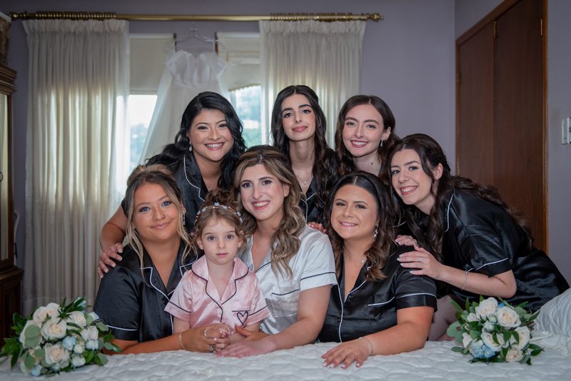 A bride, bridesmaids, and flower girl smile on a bed with floral bouquets during pre-wedding preparations.