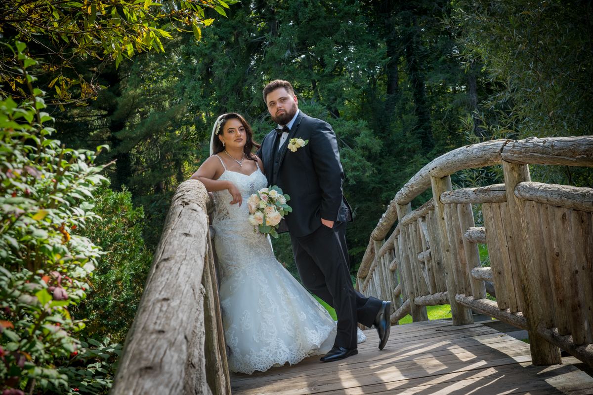 A bride and groom stand on a rustic wooden bridge surrounded by lush greenery, sunlight filtering through trees.