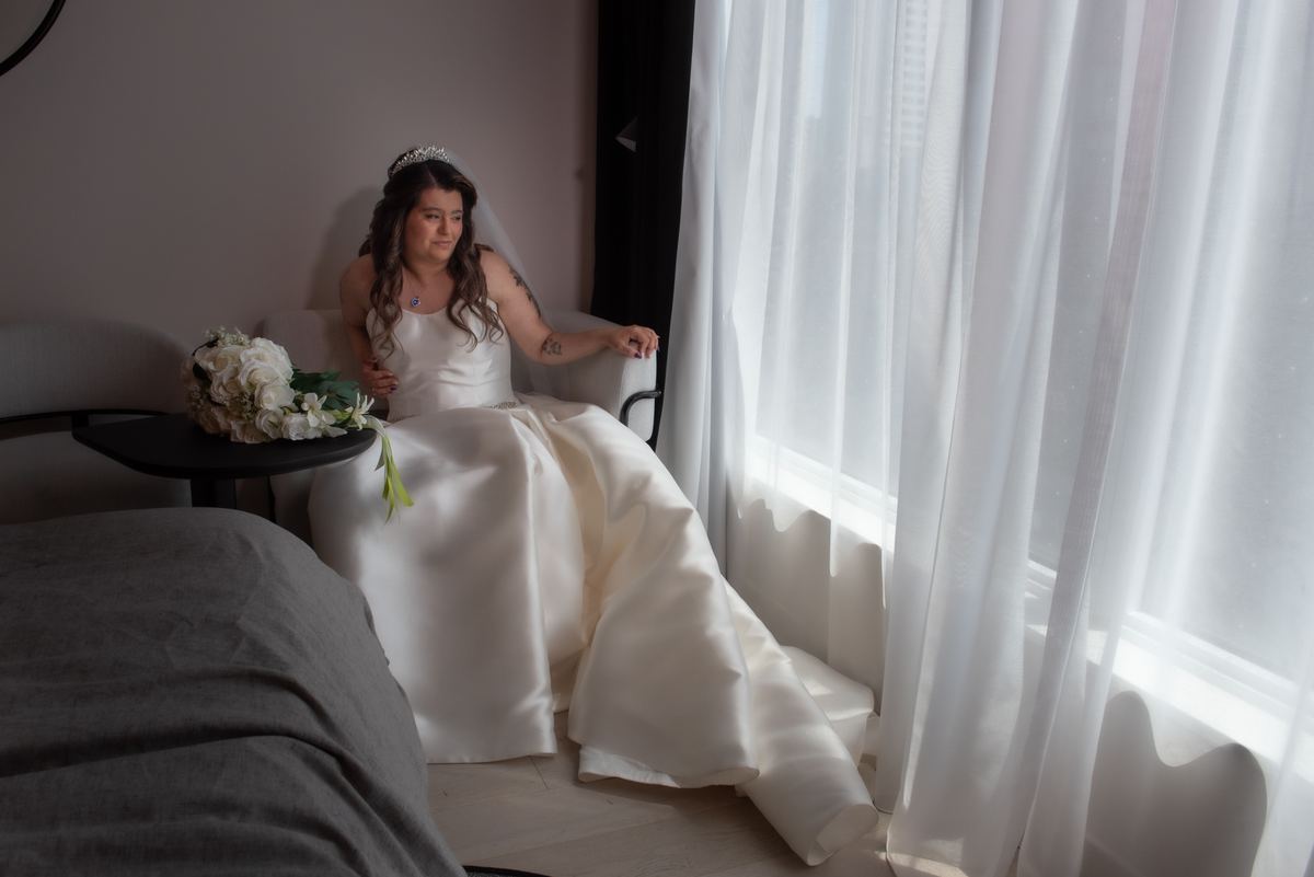 A bride in a satin gown sits by a window, gazing thoughtfully as sunlight filters through sheer curtains.