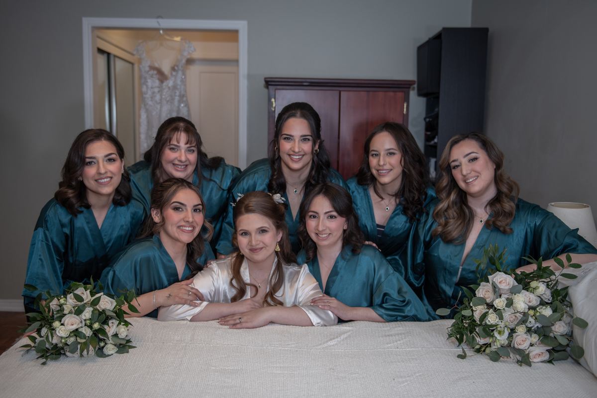 A bride and her bridesmaids are gathered on a bed in a softly lit room, smiling for a pre-ceremony portrait.