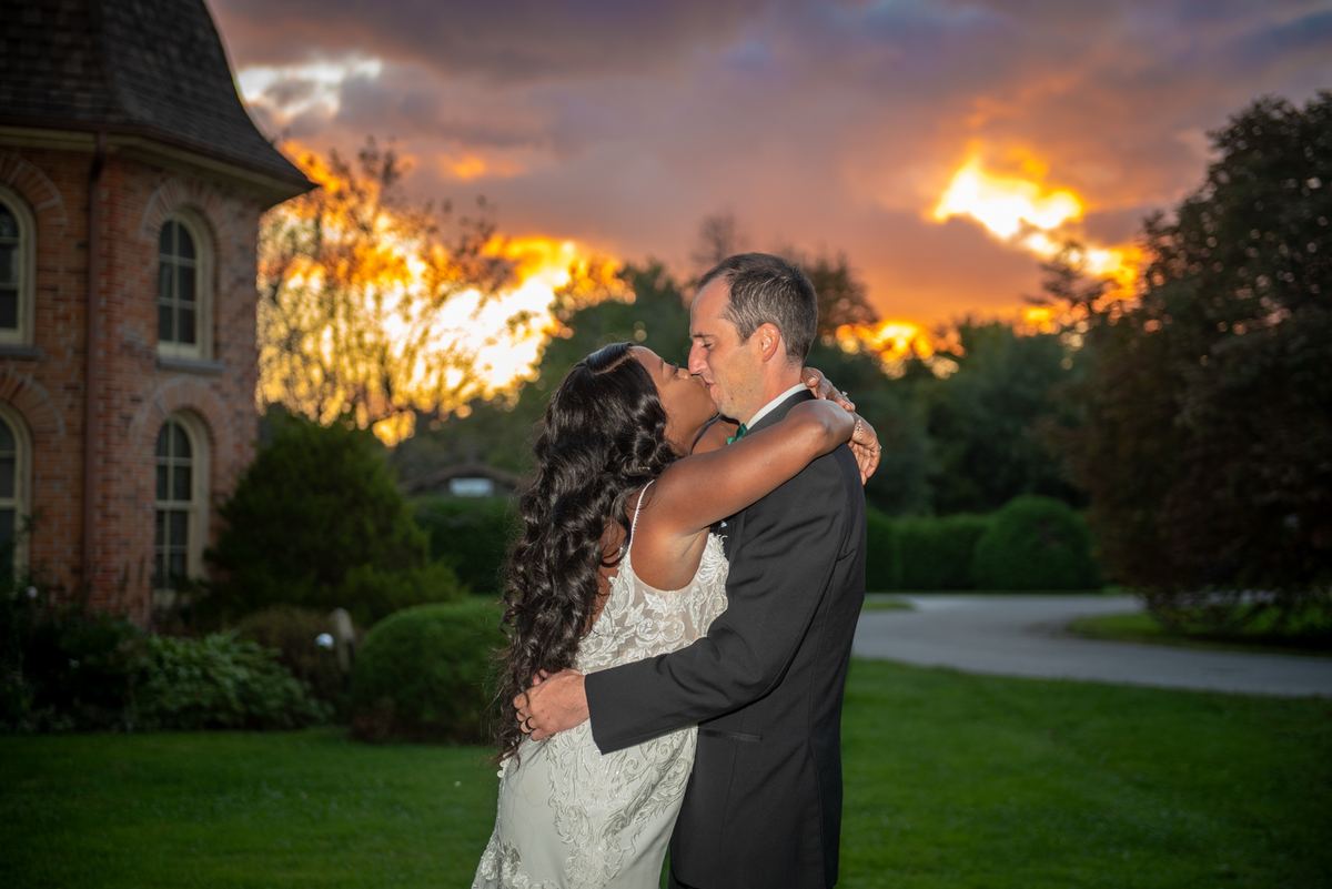 Bride and groom kiss in a garden at sunset, surrounded by orange and purple sky, trees, and a historic brick building.