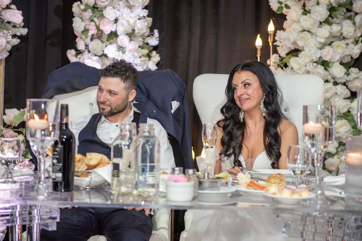 A bride and groom smile at each other during a warmly lit, elegantly decorated wedding reception table setting.