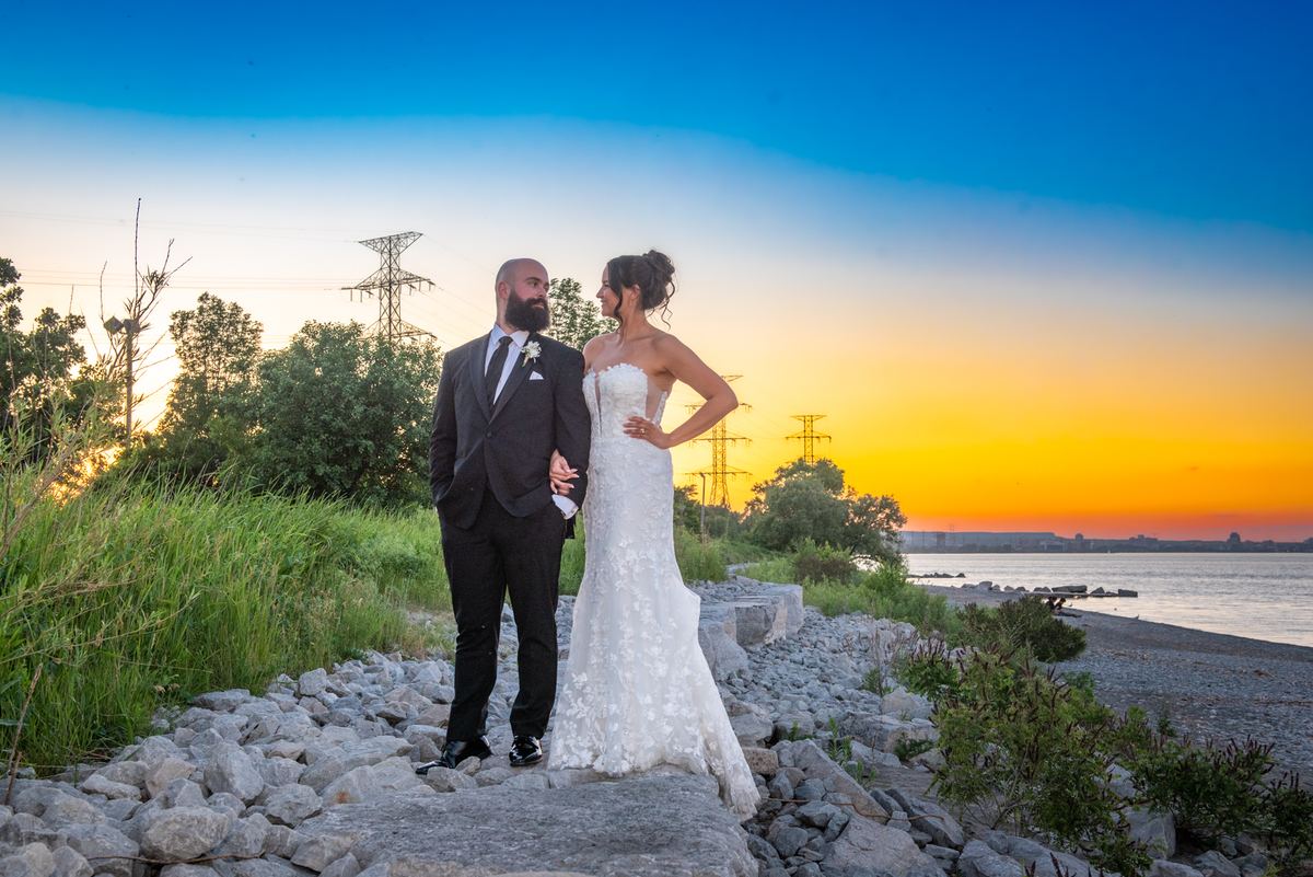 A bride in a lace gown and veil and a groom in a tuxedo stand on a rocky shoreline, facing each other in golden sunset light.