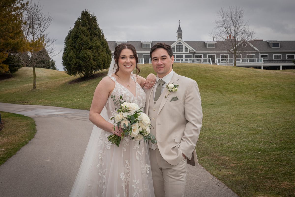 A smiling bride and groom stand on a path with a grassy hill and stately building behind them, under an overcast sky.