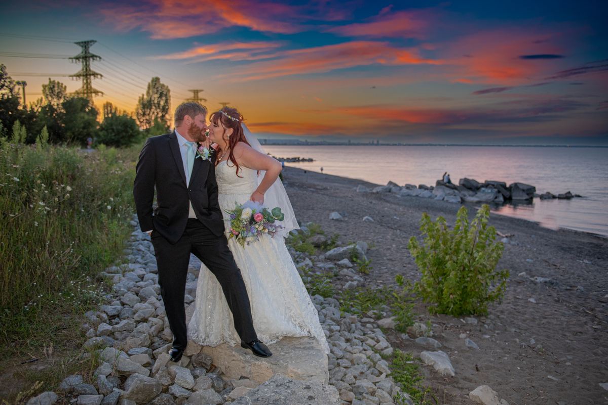 A bride and groom kiss on a rocky shore at sunset, silhouetted against a vibrant sky over the water.