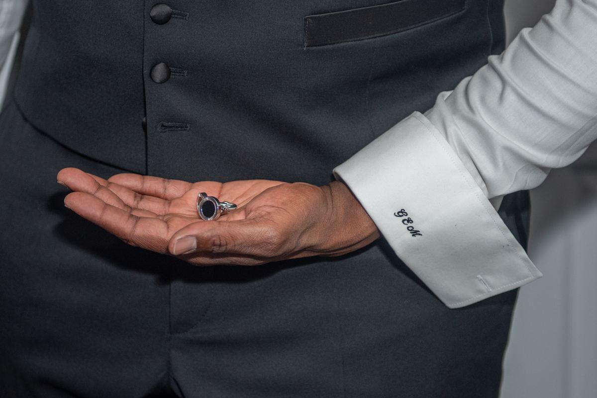 A man in formal attire presents an open palm holding a ring, illuminated softly against a muted background.
