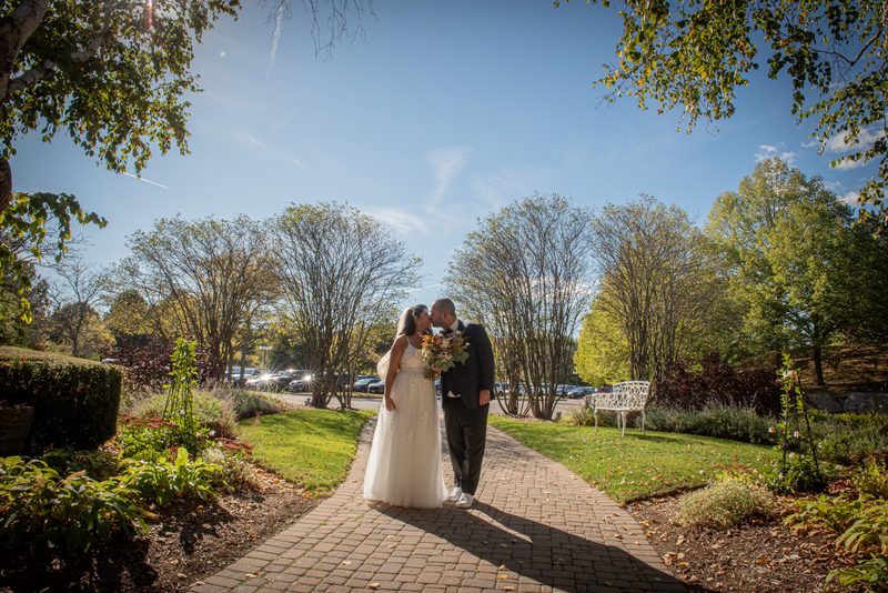 A bride and groom kiss on a sun-dappled brick pathway surrounded by autumnal trees and greenery under a clear blue sky.