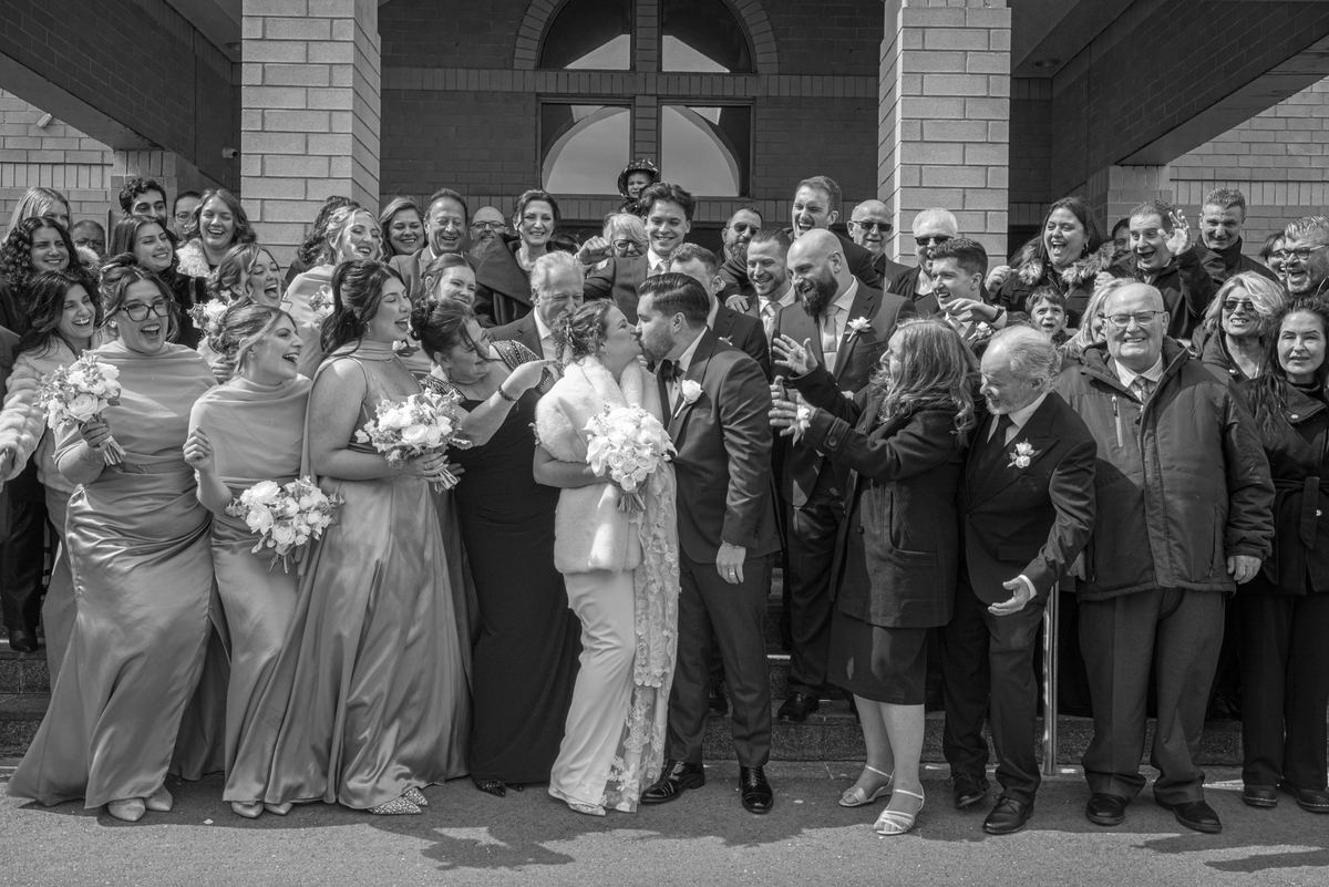 A bride and groom kiss outside with their smiling wedding party in the background, illuminated by natural light.