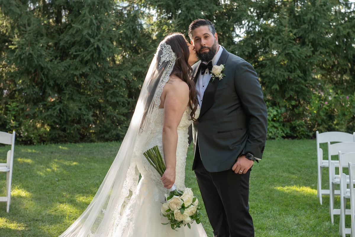 Bride in lace-trimmed veil leans to kiss groom on cheek, standing under dappled sunlight filtering through trees.