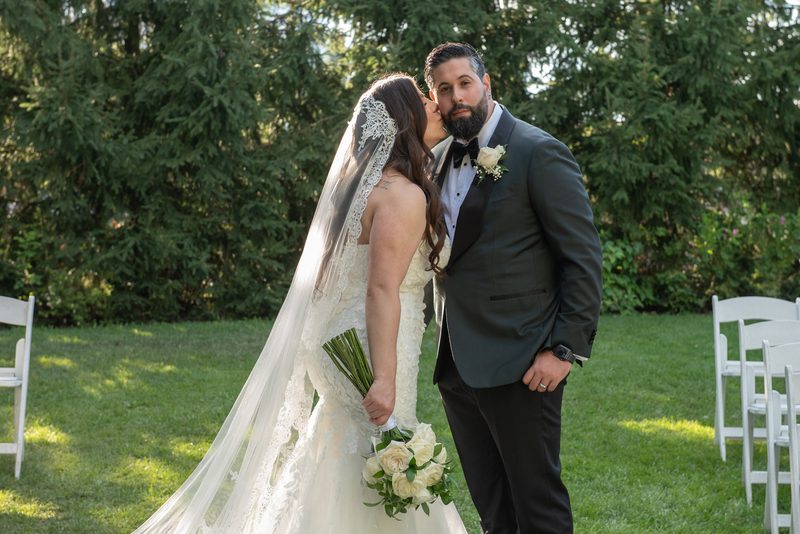 Bride in lace-trimmed veil leans to kiss groom on cheek, standing under dappled sunlight filtering through trees.