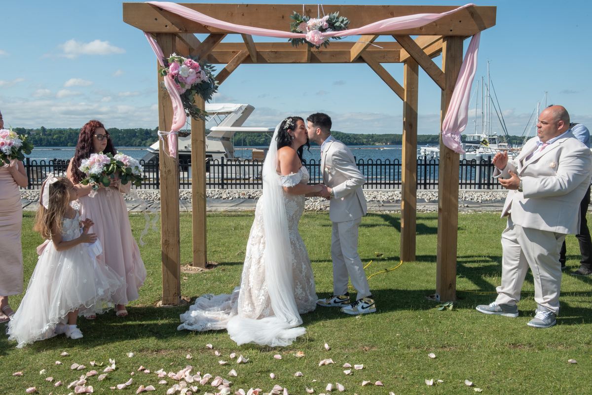 Bride and groom kiss under wooden arbor adorned with pink ribbons and flowers, surrounded by guests on grassy waterfront.