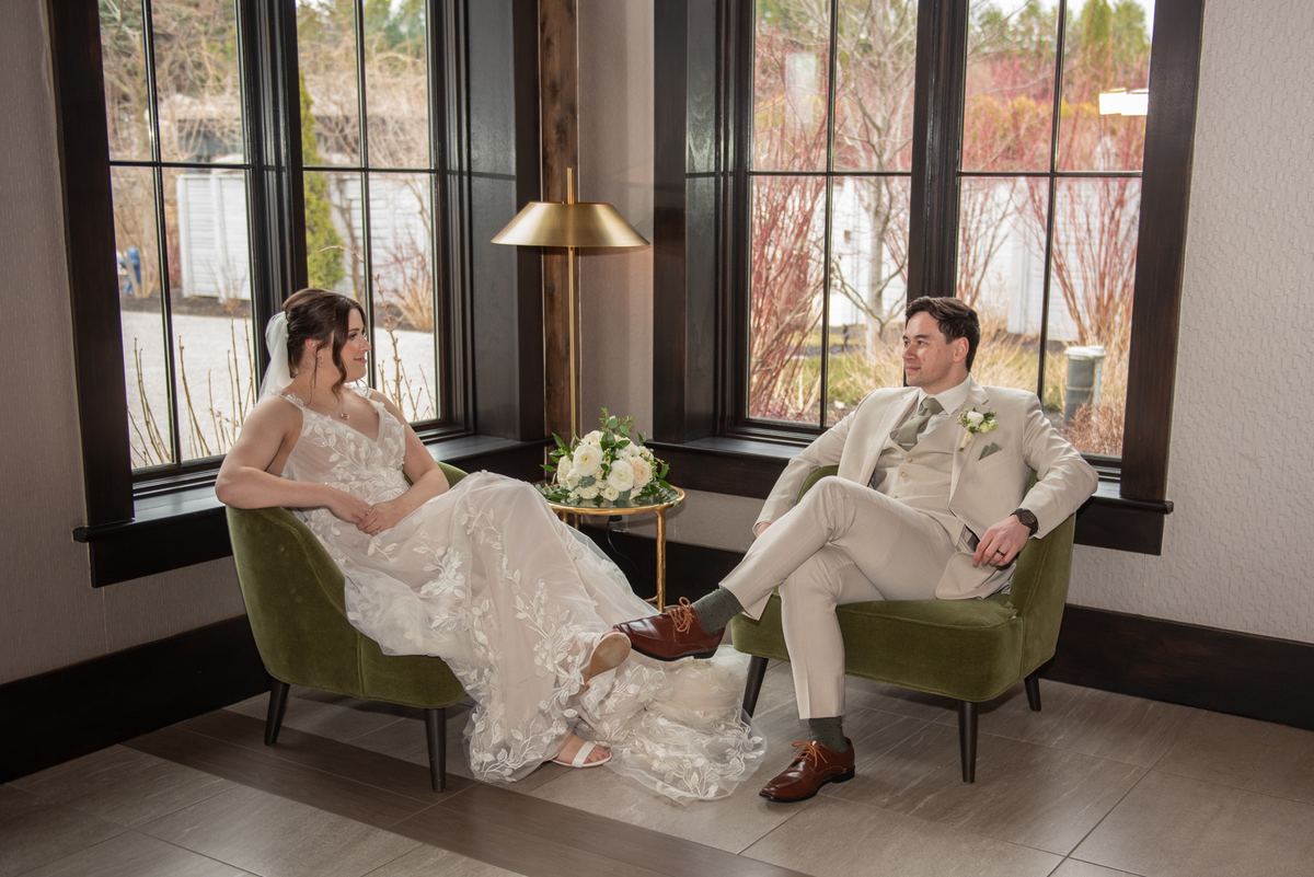 A bride and groom sit in green chairs, sharing a quiet moment with soft light filtering through windows.