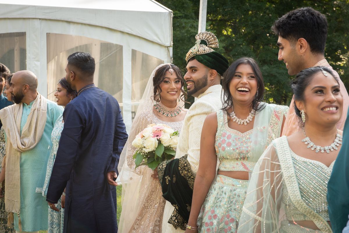 Bride and groom smile under white tent with family in traditional South Asian attire as guests laugh and celebrate.