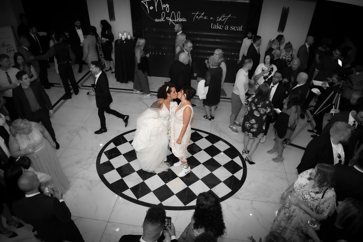 Two brides kiss at the center of a black-and-white checkered dance floor surrounded by guests holding drinks.