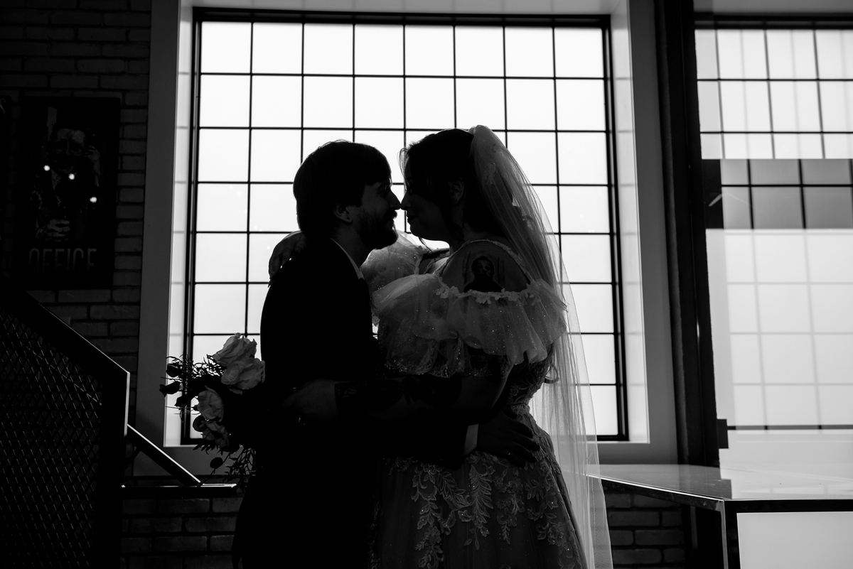 A bride and groom embrace with foreheads touching, framed by a bright window in high-contrast black-and-white.