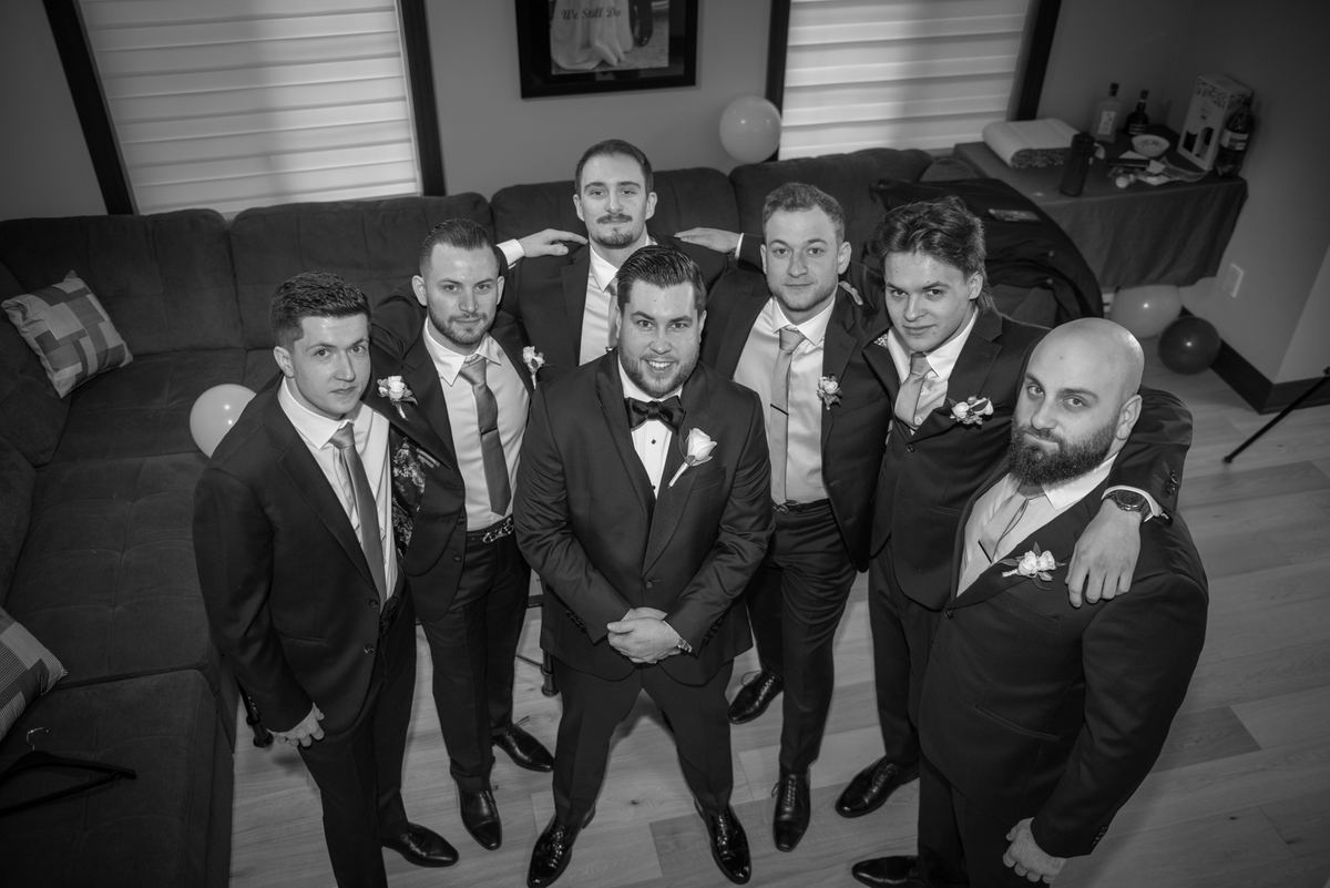 A groom and his groomsmen pose in formal suits with boutonnieres indoors, under soft diffused light from blinds.