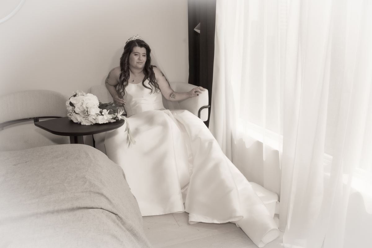 A bride in her wedding gown sits quietly by a window, gazing thoughtfully as light filters through curtains.
